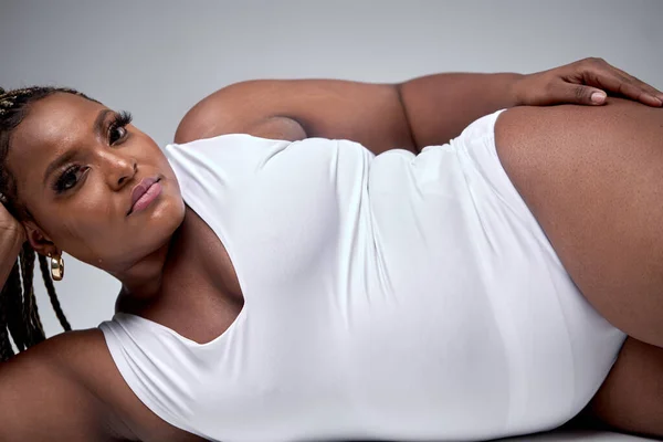 Young pretty plus size or plump woman with chubby natural body holding  napkin, posing, tired after photoshoot. beautiful female model in white  bodysuit against gray background. Lifestyle portrait Stock Photo