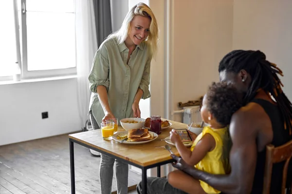 Diversa madre, padre desayunando con su hija sentada detrás de la mesa con comida —  Fotos de Stock