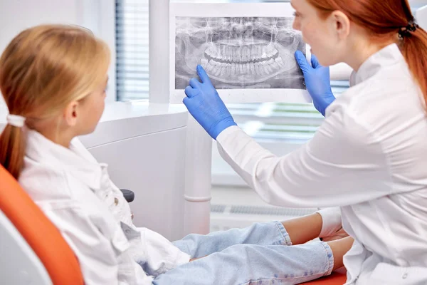 Дитина сидить у стоматологічному кріслі, дивлячись на рентгенівський знімок з лікарем, обговорює — стокове фото