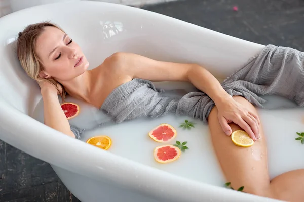 Donna bionda sexy in bagno con latte e frutta. Bagno giovanile per il corpo. — Foto Stock