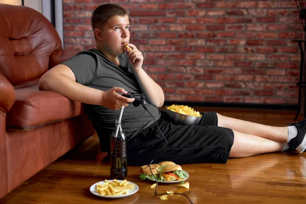 Fat boy sitting on floor in living room, side view. Overweight caucasian child at home