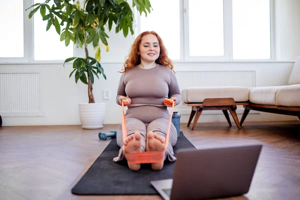 Kızıl saçlı kadın rahat oturma odasında egzersiz yaparken dizüstü bilgisayarına bakıyor.. — Stok fotoğraf