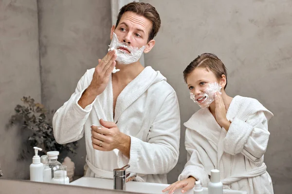 Uomo e bambino con schiuma da barba sui volti guardando nello specchio del bagno — Foto Stock