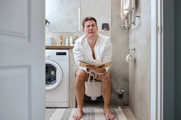 Uomo seduto in bagno sentirsi infelice e tiene libro, diarrea costipazione, concetto di salute — Foto Stock