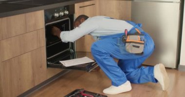 Üniformalı siyah tamirci mutfakta bozuk fırını inceliyor, notlar alıyor.