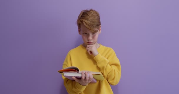 Pelirroja niño leyendo libro, pensando, de vuelta a la escuela. aislado en púrpura. — Vídeos de Stock