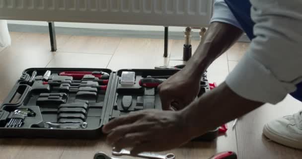 Черный профессиональный сантехник устанавливает радиатор в пустой комнате — стоковое видео