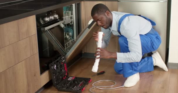 Young black plumber in overalls holding pipe while changing it in dishwasher — Vídeo de Stock