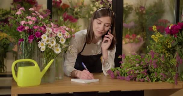 Bonita florista mulher em preto smartphone falar no telefone, ouvindo pedidos de clientes — Vídeo de Stock