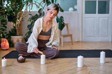woman sits meditating in lotus position on floor, using incense stick and smoke.