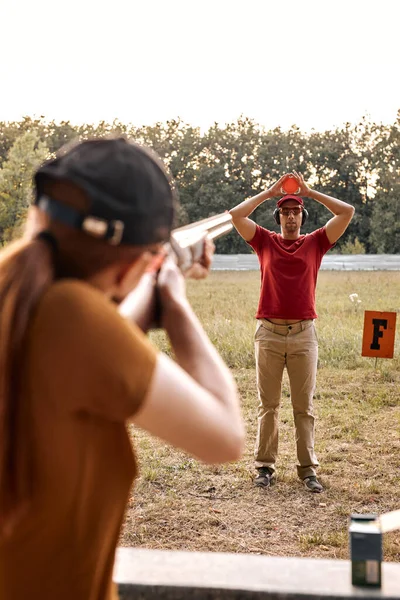 Vista trasera de la hembra en gorra y gafas apuntando rifle a la arcilla paloma objetivo en manos de macho — Foto de Stock