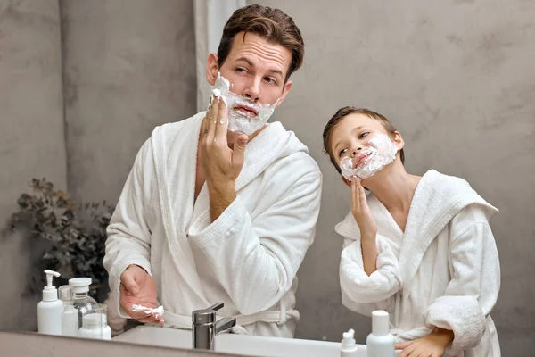 Hombre adulto y adolescente se divierten con espuma durante el afeitado juntos. Kid hijo imita padre — Foto de Stock