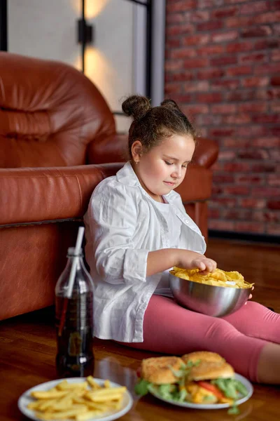 Divertente ragazza grassa mangiare patatine dalla ciotola mentre seduto sul pavimento in soggiorno, vista laterale — Foto Stock