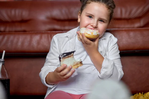 Caucasico ragazza sta mangiando malsano dolci torte e guardare la tv seduto sul pavimento. — Foto Stock
