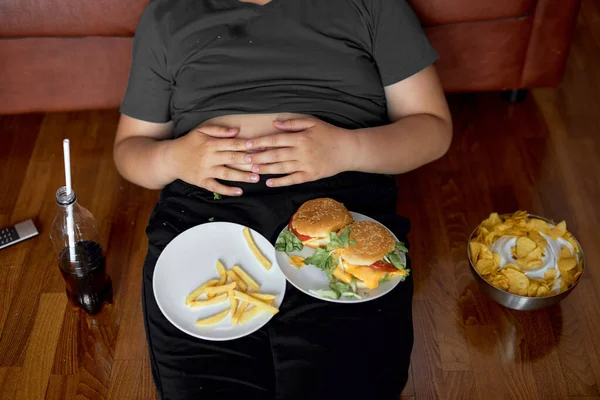 Gehakt vet zwaarlijvige jongen zitten op de vloer met junk food op bord, frietjes en hamburgers — Stockfoto
