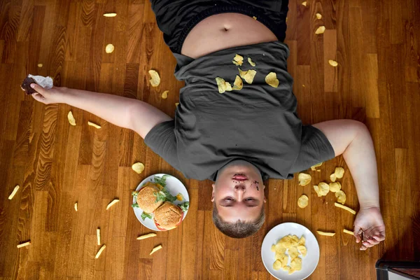 Grasso ragazzo corpulento mangiare cibo spazzatura mentre sdraiato sul pavimento a casa, con patatine fritte — Foto Stock