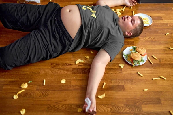 Overweight boy sleeping on floor surrounded by fast food, overeating — Stok fotoğraf