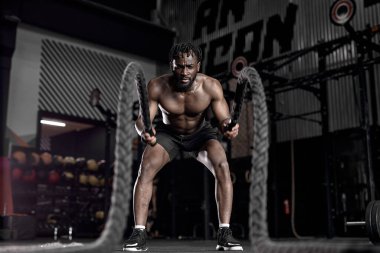 Portrait of strong young african man exercising with battle ropes during workout
