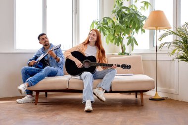 Couple hold guitar and laugh sitting together in living room. Rest time, music concept
