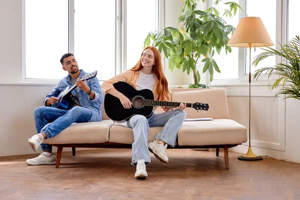 Пара держат гитару и смеются сидя вместе в гостиной. Отдых, музыкальная концепция — стоковое фото