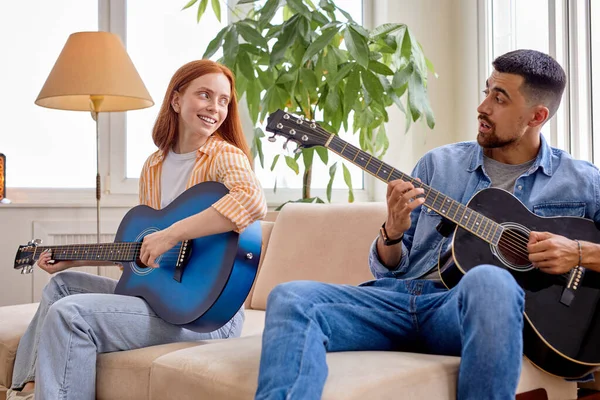 Приятная приятная женщина, сидящая на удобном диване, играющая на гитаре, учится проводить время с мужчиной — стоковое фото