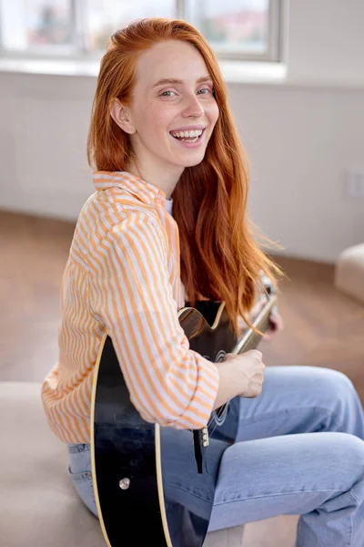 Joyeuse dame rousse joyeuse assise avec guitare, sur le canapé, souriante, regardant la caméra — Photo