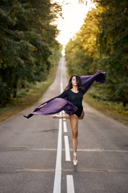 Siyah elbiseli bir kadın ve asfalt üzerinde dans eden bale ayakkabıları, zıplıyor ve eğiliyor.