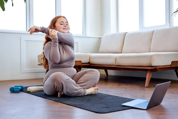 穿着运动服的红头发女人在瑜珈前张开双臂坐在健身垫上 — 图库照片
