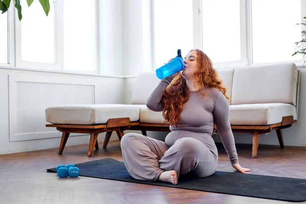 Artı kızıl saçlı kadın spor paspasının üstünde su içiyor, evde eğitim görüyor. — Stok fotoğraf