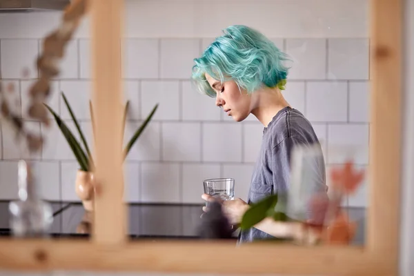 Young woman in casual clothes stand looking down in kitchen, with glass of water — Stockfoto