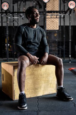 Olumlu gülen Afro-Amerikan adam çapraz oturarak ahşap kutuya atlayıp rahatlıyor.
