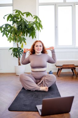 İyi görünümlü şişman kadın spor yapmak için lastik bantlarla kollarını esnetiyor, spor paspasına oturuyor.