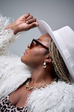 Makyajlı tombul siyah bayan gri arka planda poz veren beyaz tüylü bir ceket giyiyor.