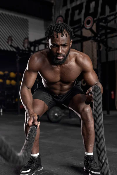 Genç Afrikalı bir adam modern karanlık spor salonunda savaş halatları kullanarak güç antrenmanı yapıyor..