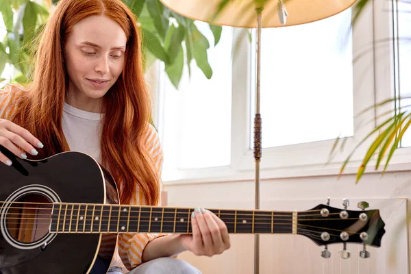 Великолепная леди с естественными рыжими волосами в повседневной одежде играет на акустической гитаре, дома — стоковое фото