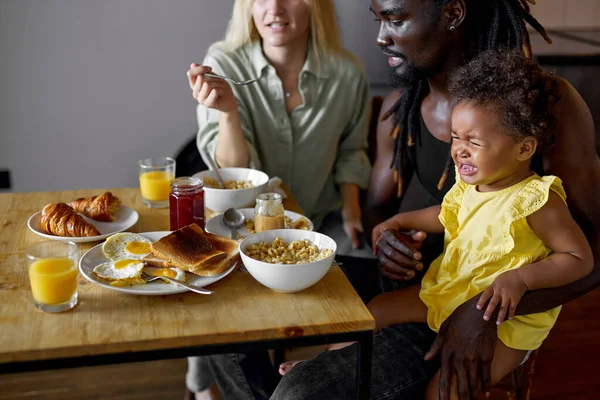 Lindo niño está llorando mientras toma el desayuno con los padres, hija traviesa —  Fotos de Stock