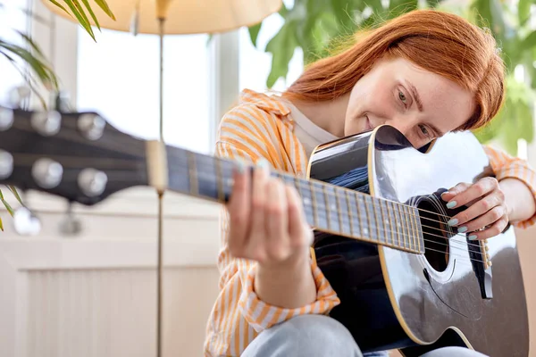 Porträtt av ung rödhårig kvinna i vardagligt slitage spela gitarr, hemma Stockbild