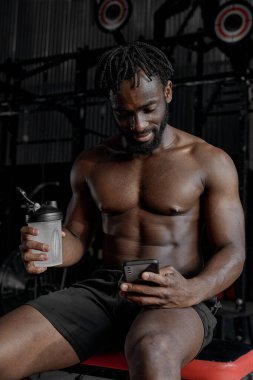 Yakışıklı sporcu, spor yaptıktan sonra dinleniyor, akıllı telefon kullanıyor, su içiyor.