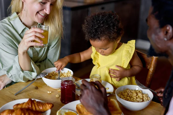 Kis fekete baba ül a etetőszékben, és eszik ételt a szülőkkel otthon. — Stock Fotó