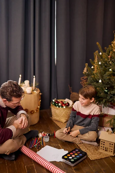 Хороший папа и сын готовятся к рождественскому празднику. рисунок на полу возле рождественской елки — стоковое фото