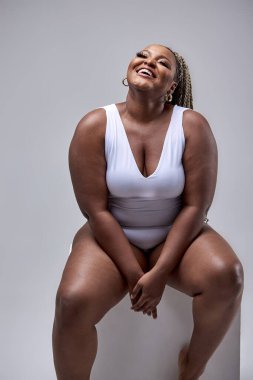 Şişkin, iri yapılı, vücut kıyafetli zenci bir kadın gülüyor, stüdyoda beyazlar içinde oturuyor.