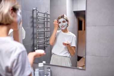 Güzel genç bir bayan aynaya bakar banyoda, evde yüzüne krem sürer.