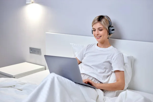 Счастливая женщина позирует с музыкой в наушниках, используя ноутбук, сидя на кровати, пишет сообщения — стоковое фото