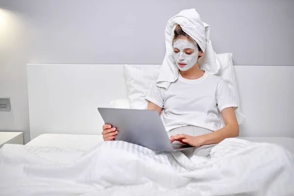 샤워 후젊고 아름다운 여자, 진흙 마스크를 쓰고 침대에서 노트북으로 타자를 치는 모습 — 스톡 사진