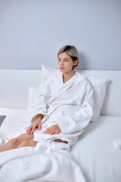 Short haired woman in bathrobe sitting on bed at home, relaxing, having rest — Stock Photo, Image