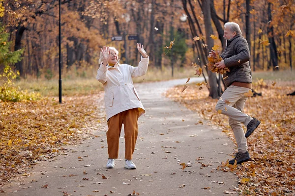 Radostný starší pár baví společně v parku při chůzi, muž zvrací listí — Stock fotografie