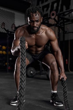 Genç siyahi bir adam modern karanlık spor salonunda savaş halatları kullanarak antrenman yapıyor..