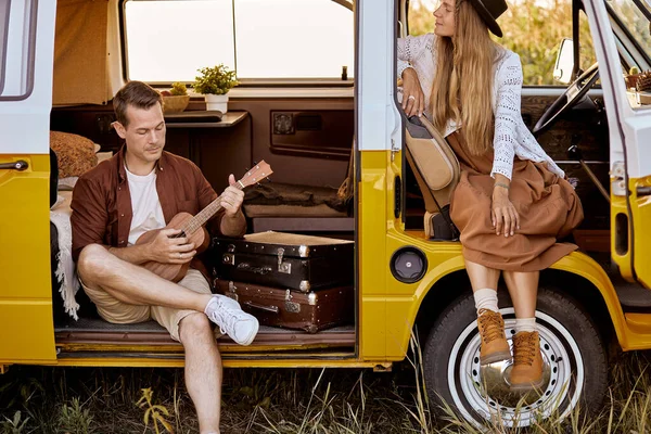 Caucasico hippie coppia sorridente, giocando ukulele in retro minivan in campo — Foto Stock