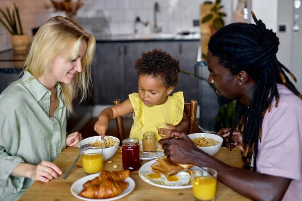 Little black child girl sitting in high chair and eating food with parents indoors. — Stock Photo, Image