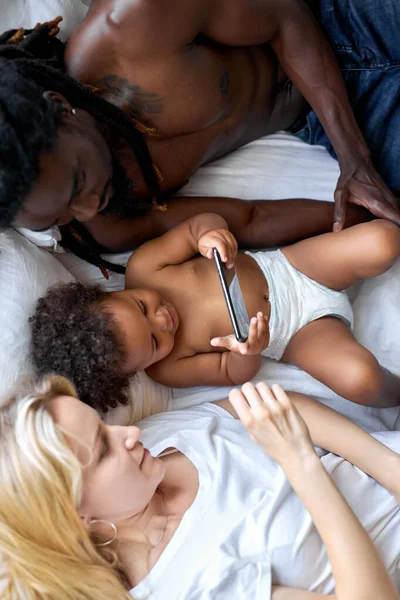 Vista superior de la familia multiétnica que descansa en la cama usando Smartphone, teléfono móvil — Foto de Stock
